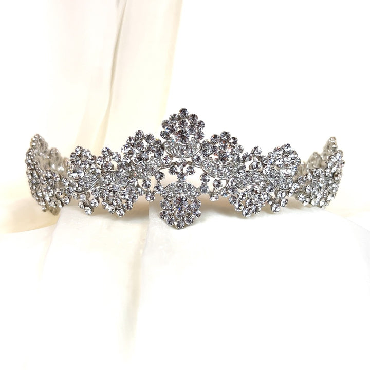 The Crystal Wedding Tiara style no. 80039, reminiscent of a Swarovski crown, features intricate floral and leaf designs adorned with numerous small, clear crystals. It stands upright on a softly blurred white background, highlighting its shimmering details.
