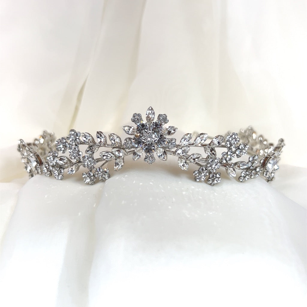 A delicate Swarovski Crystal Floral Tiara (style no. 80008, featuring sparkling floral and leaf patterns set against a soft, white fabric background.