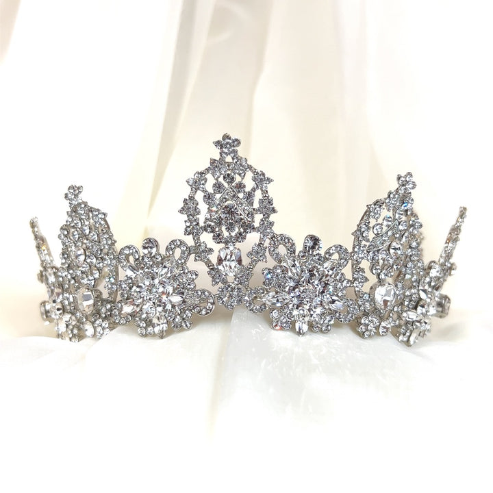 The Regal Bridal Crown style no. 80038, crafted in silver with intricate floral motifs and sparkling Swarovski crystals, is set against a soft white fabric background.