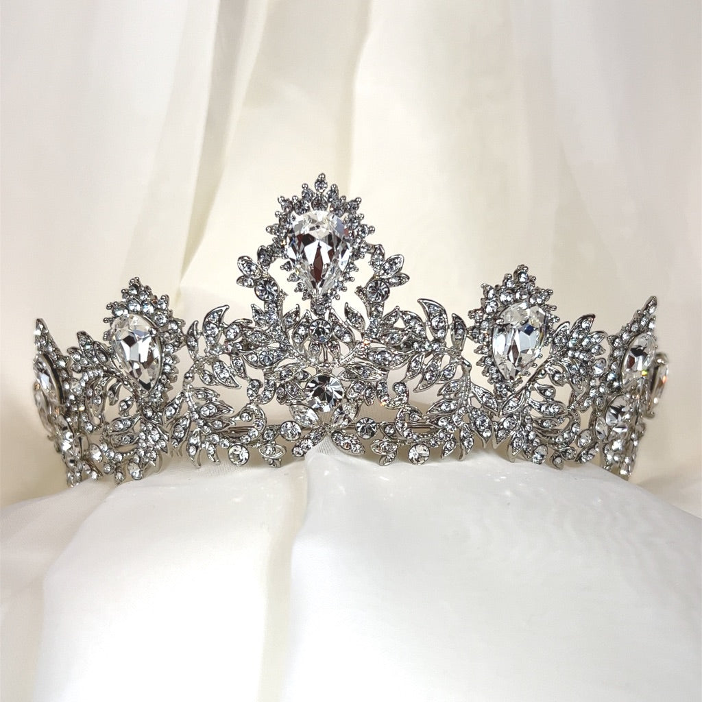 The Glamorous Swarovski Crystal Bridal Tiara style no. 80100 features intricate floral designs with sparkling crystals and is displayed against a soft white fabric backdrop.