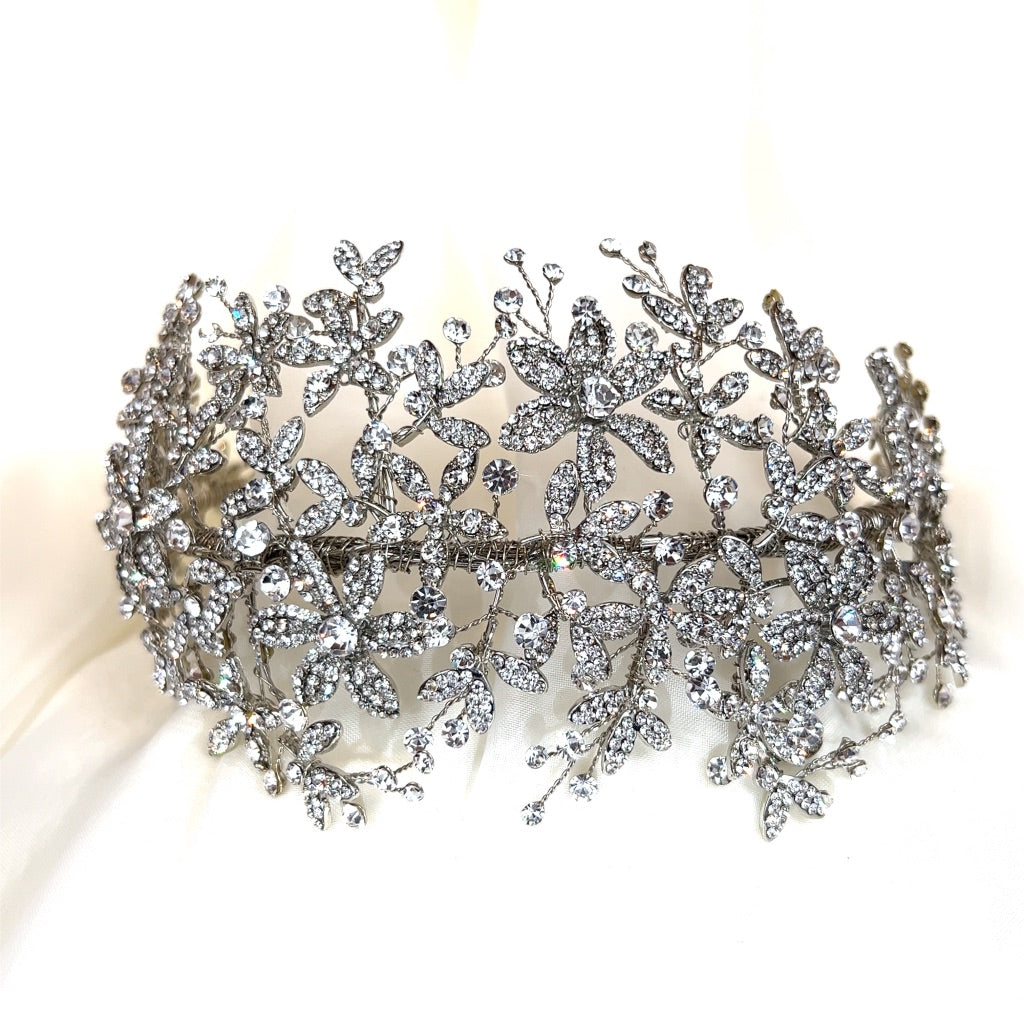 A rhodium plated Wide Floral Crystal Headband (style no. 242689) features intricate floral and leaf designs with shimmering rhinestones on a reflective surface.