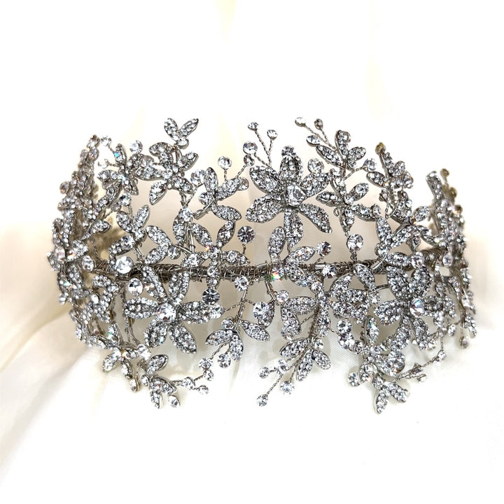 A rhodium plated Wide Floral Crystal Headband (style no. 242689) features intricate floral and leaf designs with shimmering rhinestones on a reflective surface.
