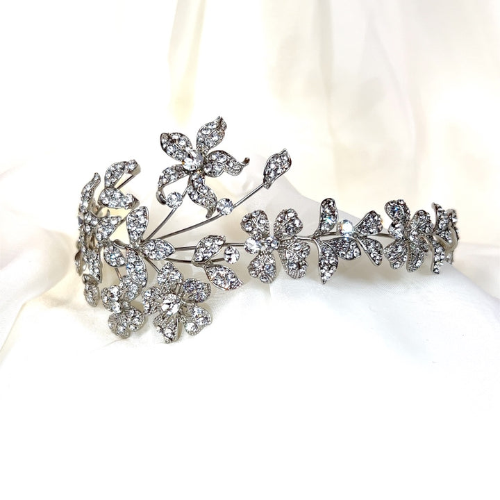 An asymmetrical floral headband, style no. 242687, featuring intricately designed flowers encrusted with Swarovski crystal diamonds is displayed on a soft white fabric background.