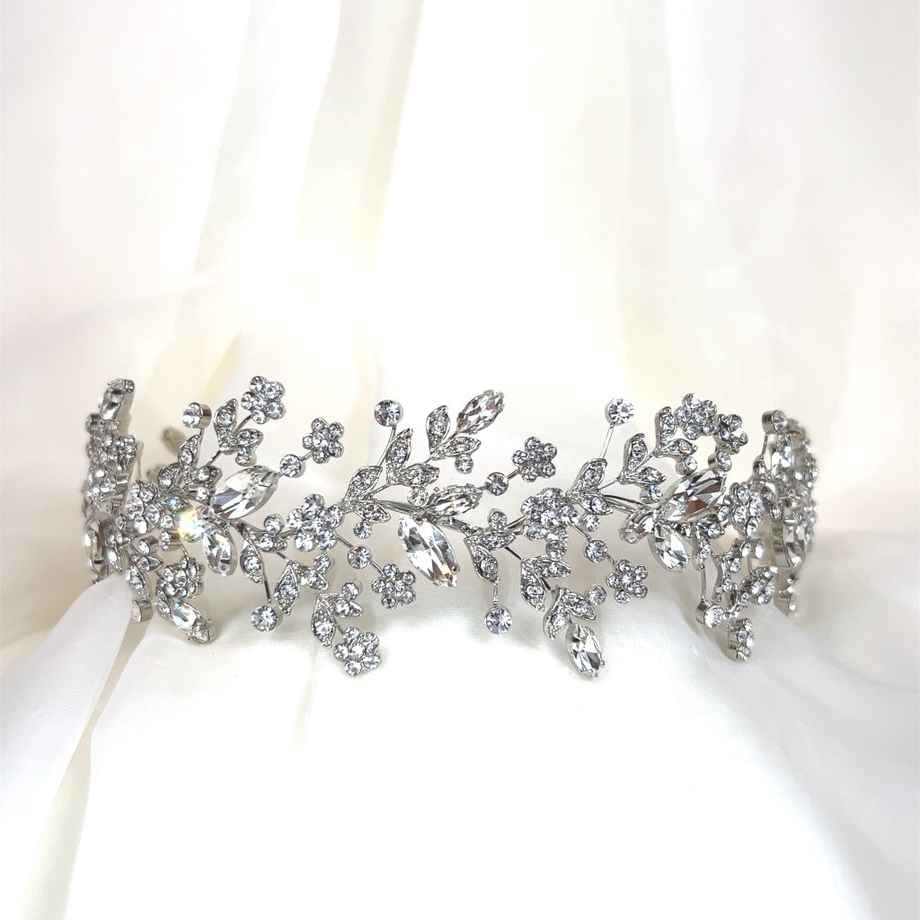 The Dainty Crystal Bridal Headband (style no. 242684), featuring intricate silver floral and Swarovski crystal leaf designs, is elegantly showcased against a soft white fabric backdrop.