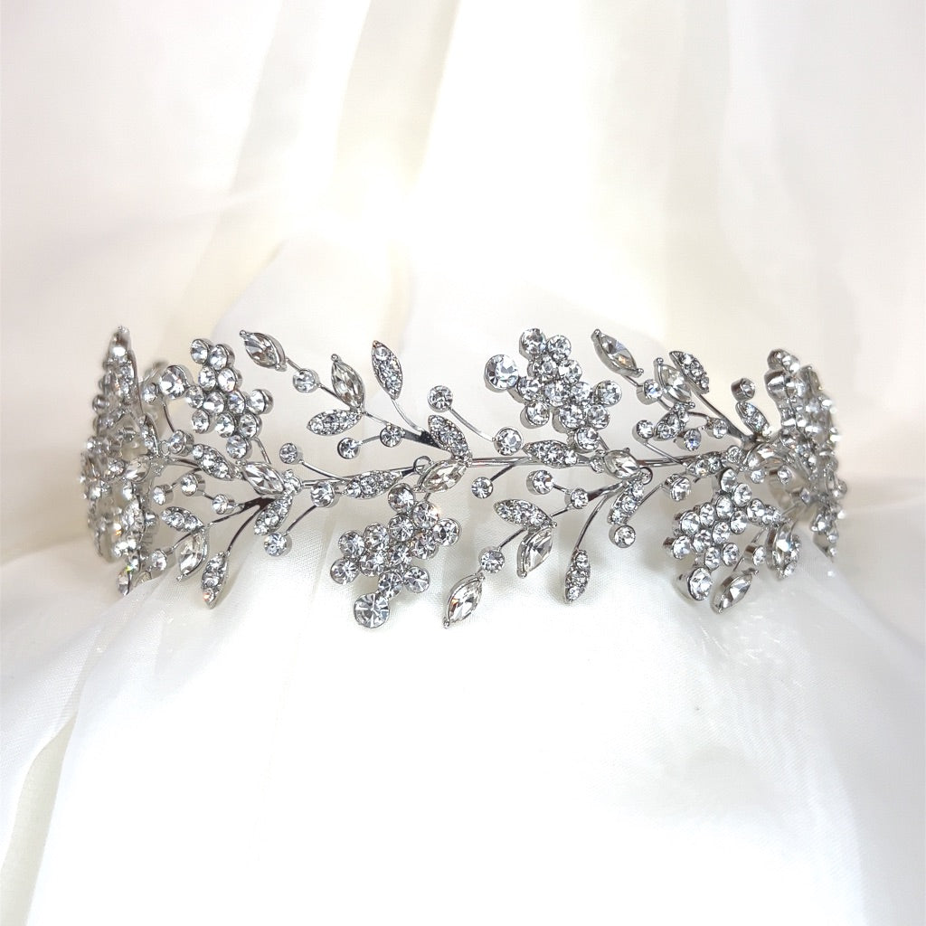 The Shimmering Cluster Headband, style no. 242685, a silver Swarovski bridal headpiece featuring intricate floral and leafy patterns encrusted with numerous sparkling crystals. It is showcased against a soft white fabric background.