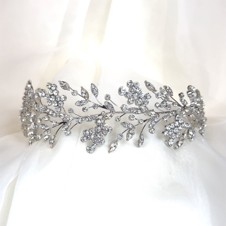 The Shimmering Cluster Headband, style no. 242685, a silver Swarovski bridal headpiece featuring intricate floral and leafy patterns encrusted with numerous sparkling crystals. It is showcased against a soft white fabric background.