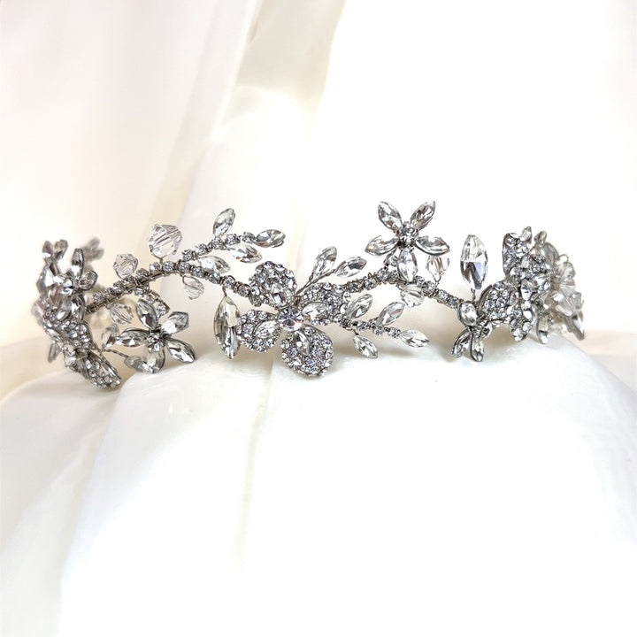 The Delicate Floral Headband, style no. 242694, with intricate gemstone patterns, is showcased next to a Swarovski bridal tiara on a soft white fabric backdrop.