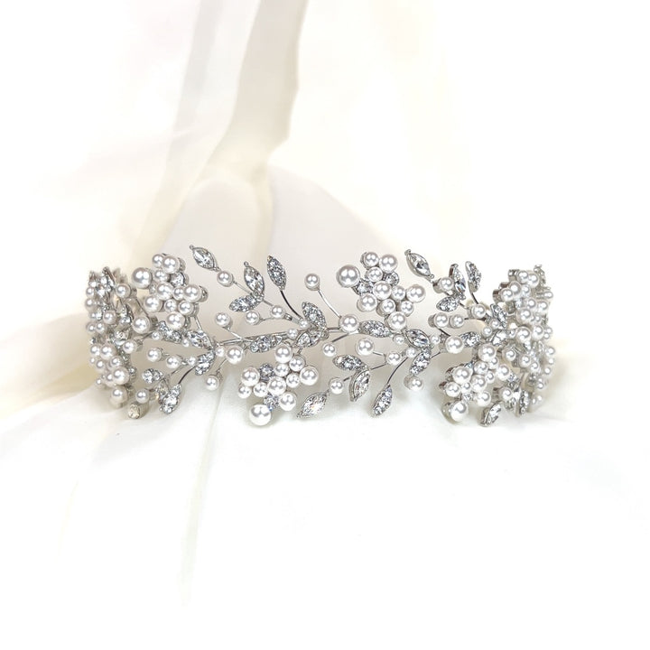 The Shimmering Cluster Headband (style no. 242685) is a delicate, dazzling Swarovski crystal bridal headpiece  featuring intricate branches of pearls and sparkling leaves, beautifully set against a soft cream-colored fabric background.