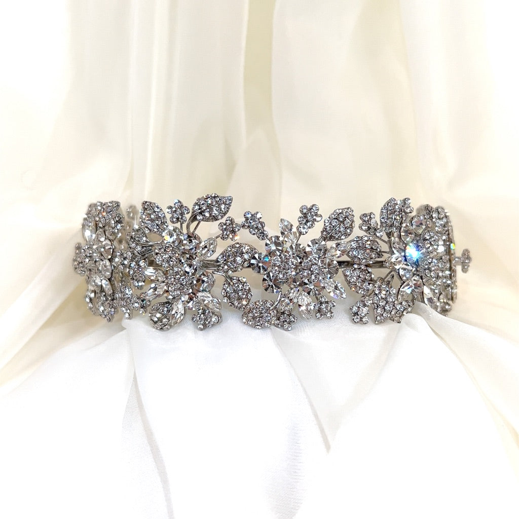 A Swarovski crystal wedding accessory with floral and leaf designs, adorned with clear gemstones, rests on white fabric.