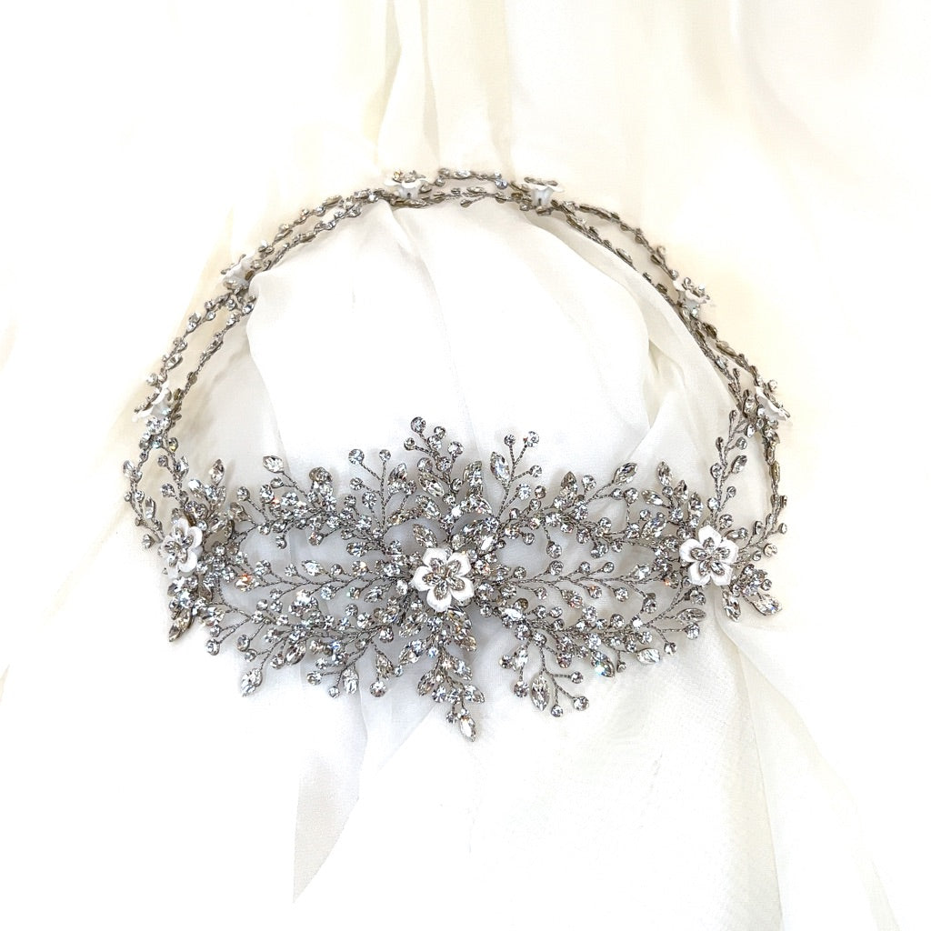 The Enchanting Swarovski Crystal and Flower Wedding Halo, style no. 242713, is a delicate crystal bridal headpiece featuring intricate floral patterns. It's beautifully set against a soft white fabric background.