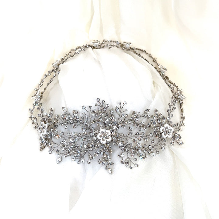 The Enchanting Swarovski Crystal and Flower Wedding Halo, style no. 242713, is a delicate crystal bridal headpiece featuring intricate floral patterns. It's beautifully set against a soft white fabric background.