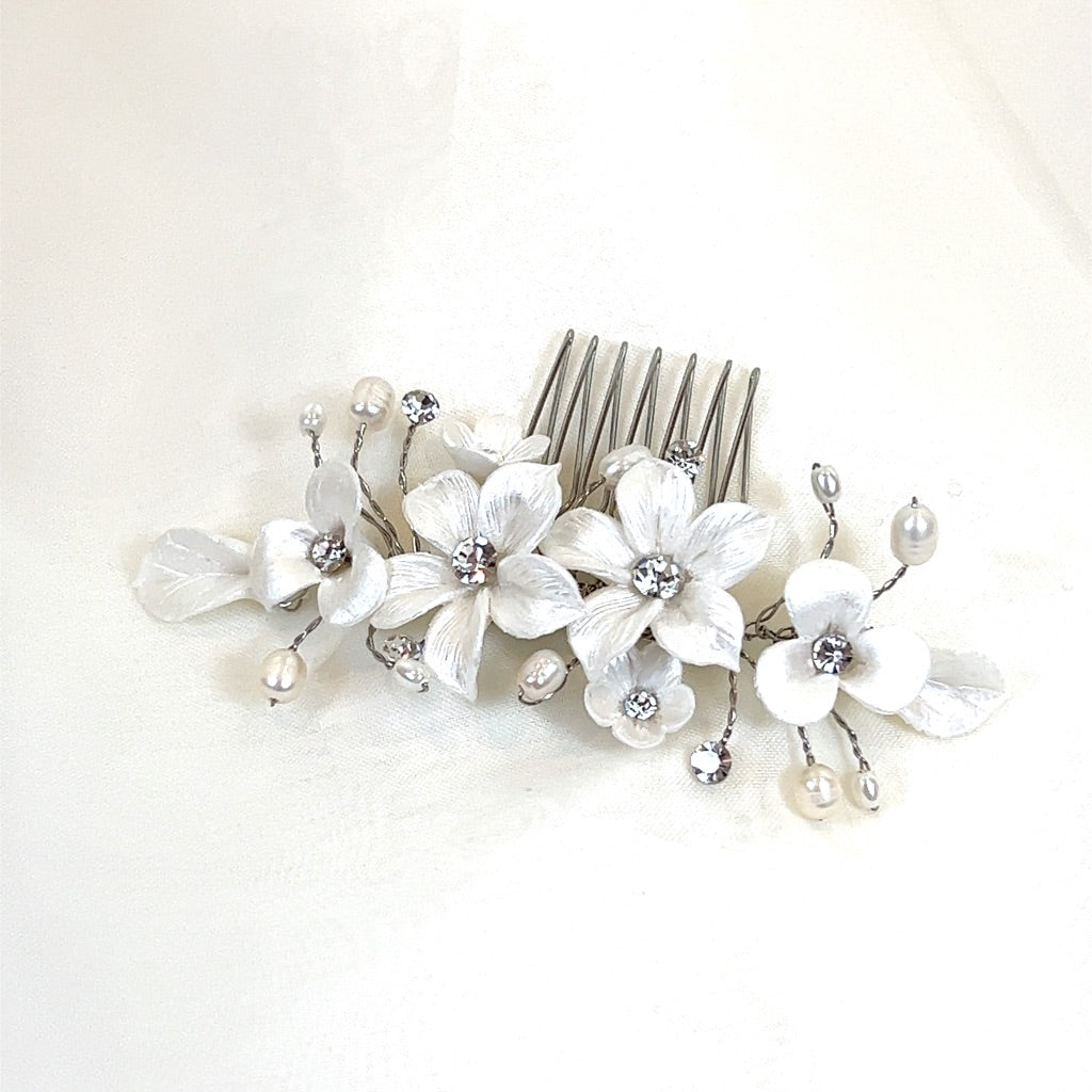 The Romantic Porcelain Flower Hair Comb (style no. 242724) is a delicate Swarovski crystal headpiece featuring dainty ceramic flowers, pearl accents, and crystals. Its elegant design with three prominent flowers and sprigs.