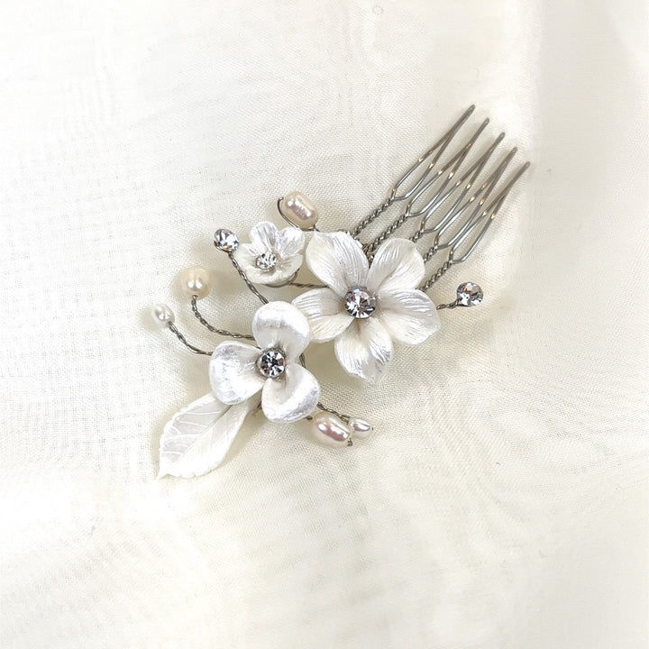 The Porcelain Flower Pin style no. 242723 is a delicate bridal hair comb adorned with white flowers, pearl accents, and sparkling Swarovski crystals against a soft cream fabric backdrop.