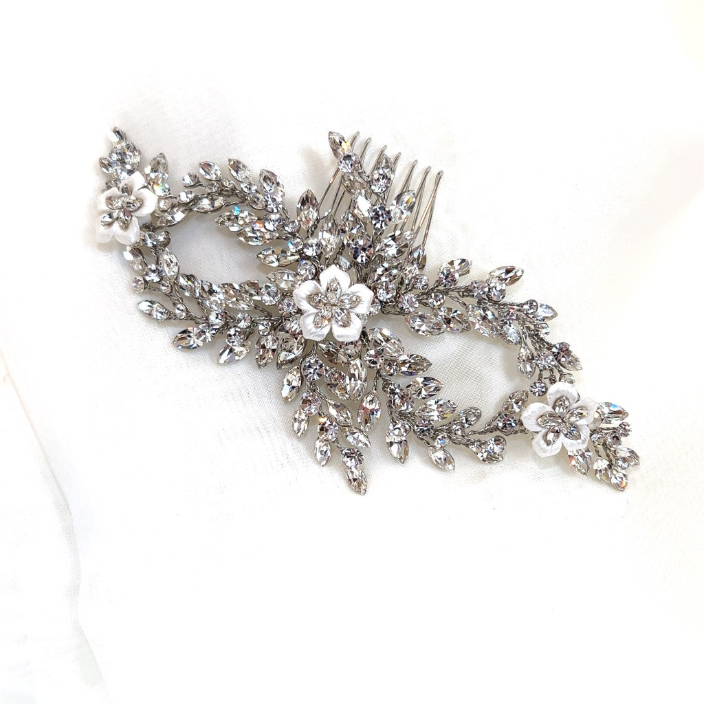 The Crystal and Porcelain Flower Bridal Comb (style no. 242726) is adorned with intricate Swarovski crystals in a leafy pattern and three white floral accents, resting on a soft white background.