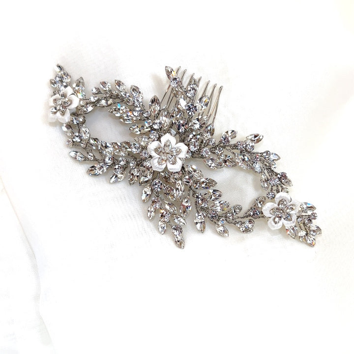 The Crystal and Porcelain Flower Bridal Comb (style no. 242726) is adorned with intricate Swarovski crystals in a leafy pattern and three white floral accents, resting on a soft white background.