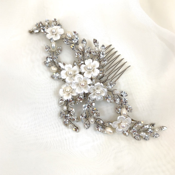 On a soft white fabric, the Looping Crystal and Flower Bridal Hair Comb (style no. 242725) features silver leaves, white flowers, and pearl accents. Covered with Swarovski crystals, pearls, and ceramic flowers, it boasts ornate elegance with intricate floral and leaf details.