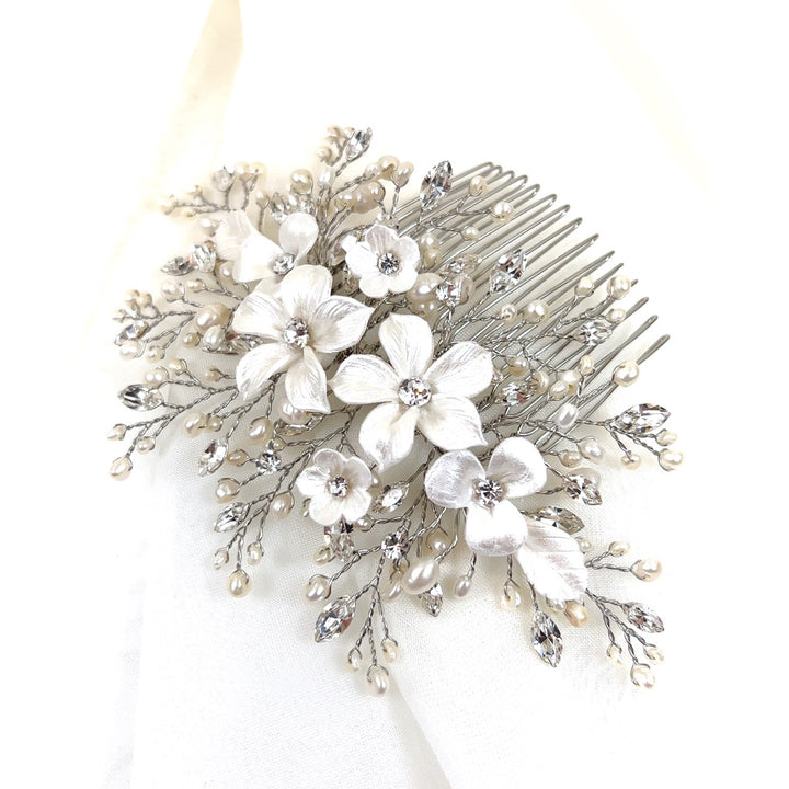 The Porcelain Flower Bouquet Wedding Hair Comb (style no. 242722) rests on a white fabric, showcasing intricate design with white ceramic flowers, pearl accents, and Swarovski crystal embellishments.