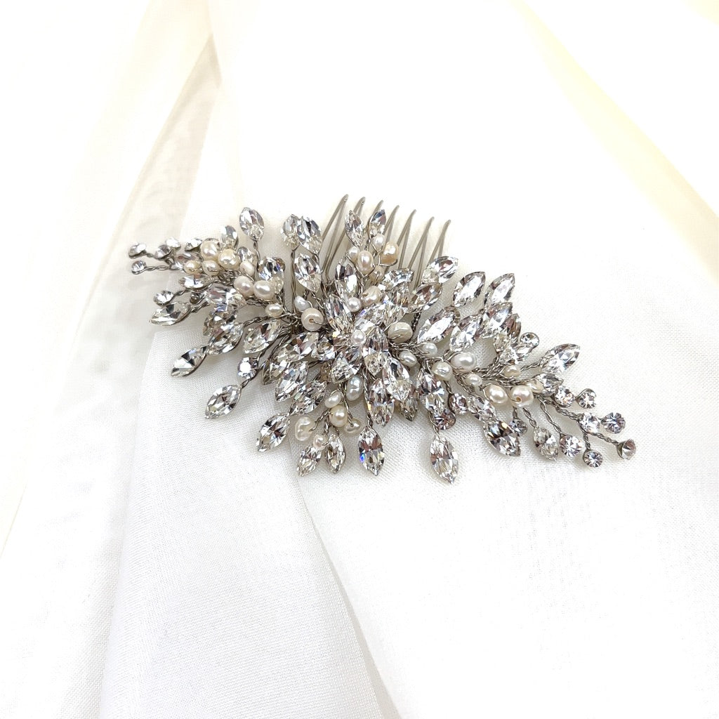 The Dainty Crystal Comb (style no. 242731) rests on a white fabric background, showcasing intricate silver details with pearl accents. Its leaf and flower clusters are adorned with sparkling Swarovski crystal jewels.