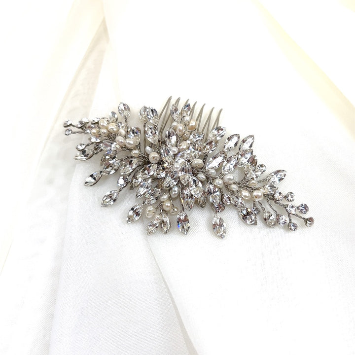 The Dainty Crystal Comb (style no. 242731) rests on a white fabric background, showcasing intricate silver details with pearl accents. Its leaf and flower clusters are adorned with sparkling Swarovski crystal jewels.