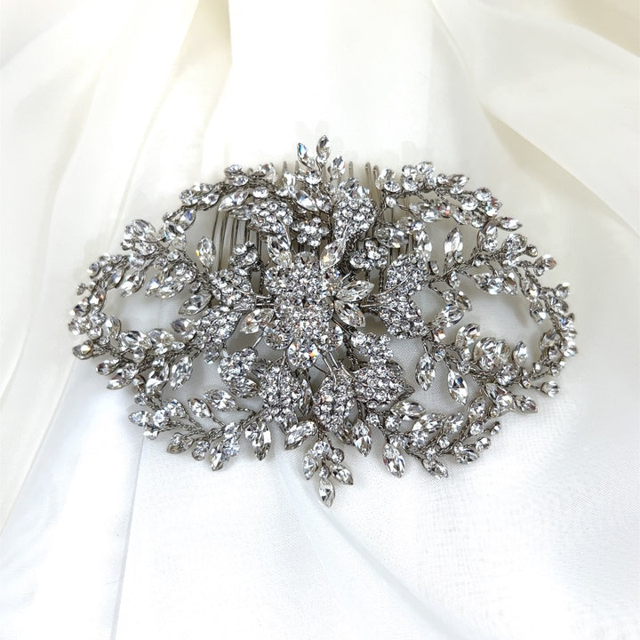 A sparkling Crystal Bloom Comb (style no. 242717) rests on white fabric, adorned with intricate floral and leaf patterns, and encrusted with shimmering Swarovski crystals that reflect light for a luxurious and elegant appearance.