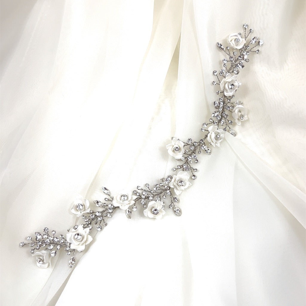 The Romantic Flower Vine, style no. 242720, is a delicate silver floral hairpiece featuring white rose details and crystal embellishments, similar to Swarovski hair combs, elegantly placed on sheer light fabric.