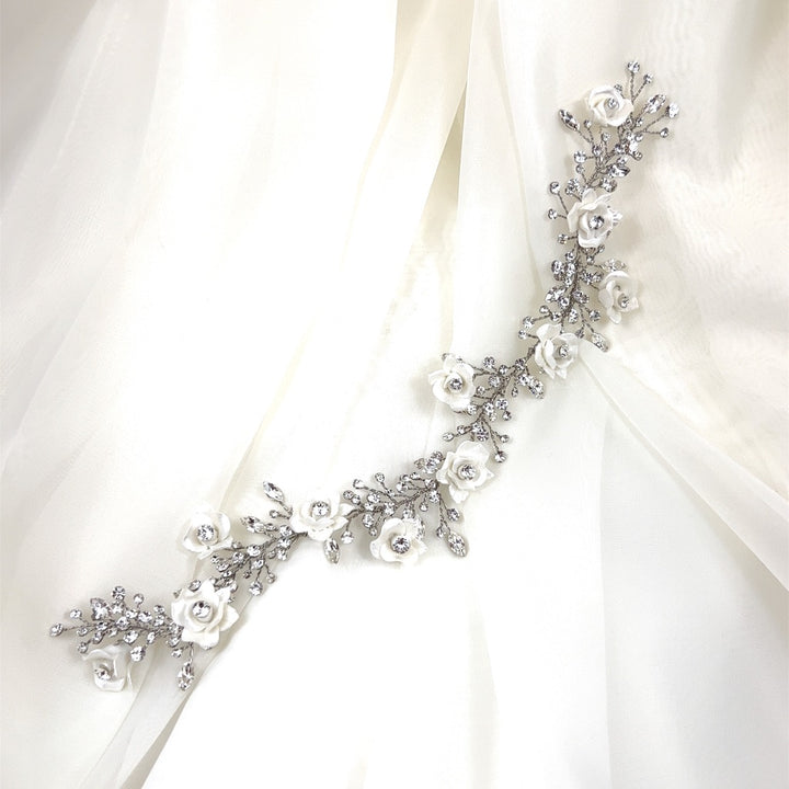 The Romantic Flower Vine, style no. 242720, is a delicate silver floral hairpiece featuring white rose details and crystal embellishments, similar to Swarovski hair combs, elegantly placed on sheer light fabric.