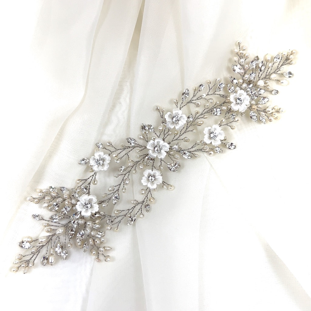 The Delicate Pearl and Porcelain Flower Vine (style no. 242712), a silver floral hairpiece featuring intricate white flowers, leaf designs, and embellished with crystals and pearls, is reminiscent of a Swarovski bridal tiara against a soft white fabric backdrop.