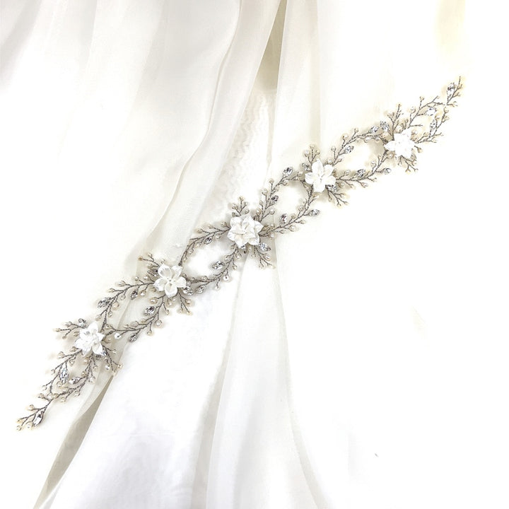 The Romantic Looping Blossom Hair Vine, style no. 242718, is an elegant Swarovski crystal hair accessory with intricate silver branches and white flowers displayed on a flowing white fabric.