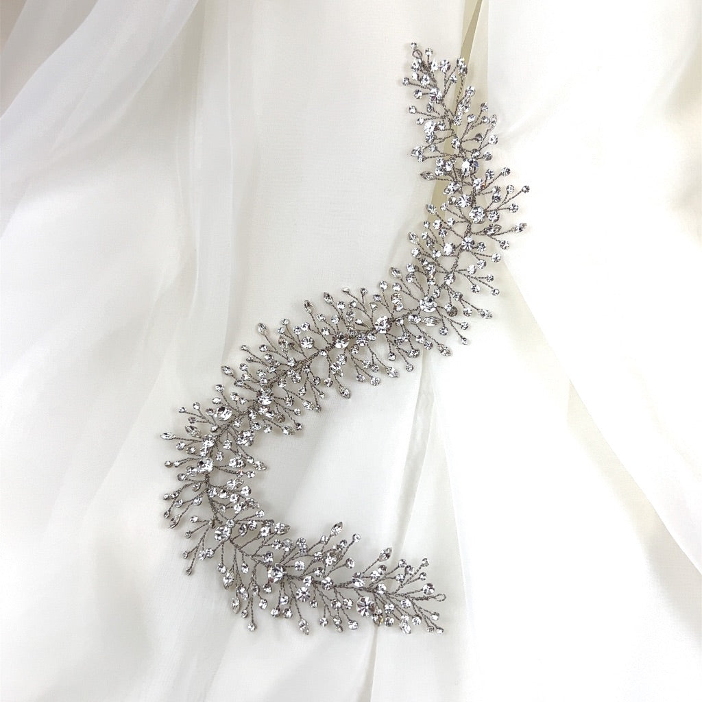 The Elegant Branching Crystal Hair Vine (style no. 242699) is a dazzling Swarovski hair vine featuring intricate leaf and crystal details, elegantly displayed on white, flowing fabric.