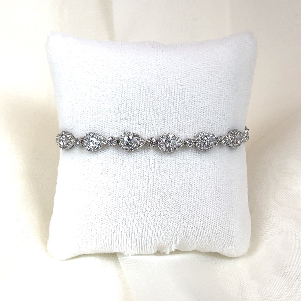 The Dainty Classic Teardrop Bracelet (style no. B146), in a silver hue, features pear-shaped and round CZ stones and is elegantly displayed on a white fabric cushion.