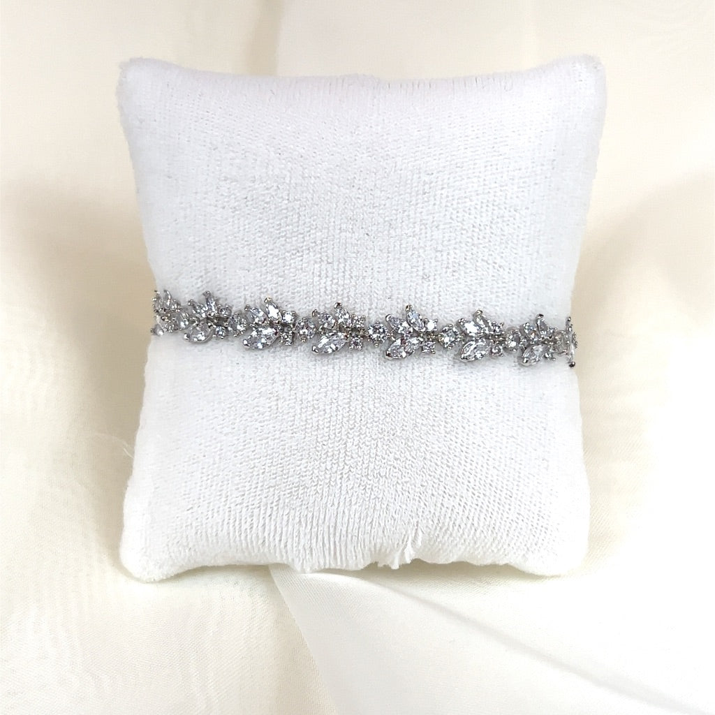 The Delicate Floral CZ Bracelet (style no. B149) is a bridal bracelet with a leaf pattern and sparkling gems, reminiscent of a Swarovski hair vine, shown on a white cushioned stand against a soft cream-colored backdrop.