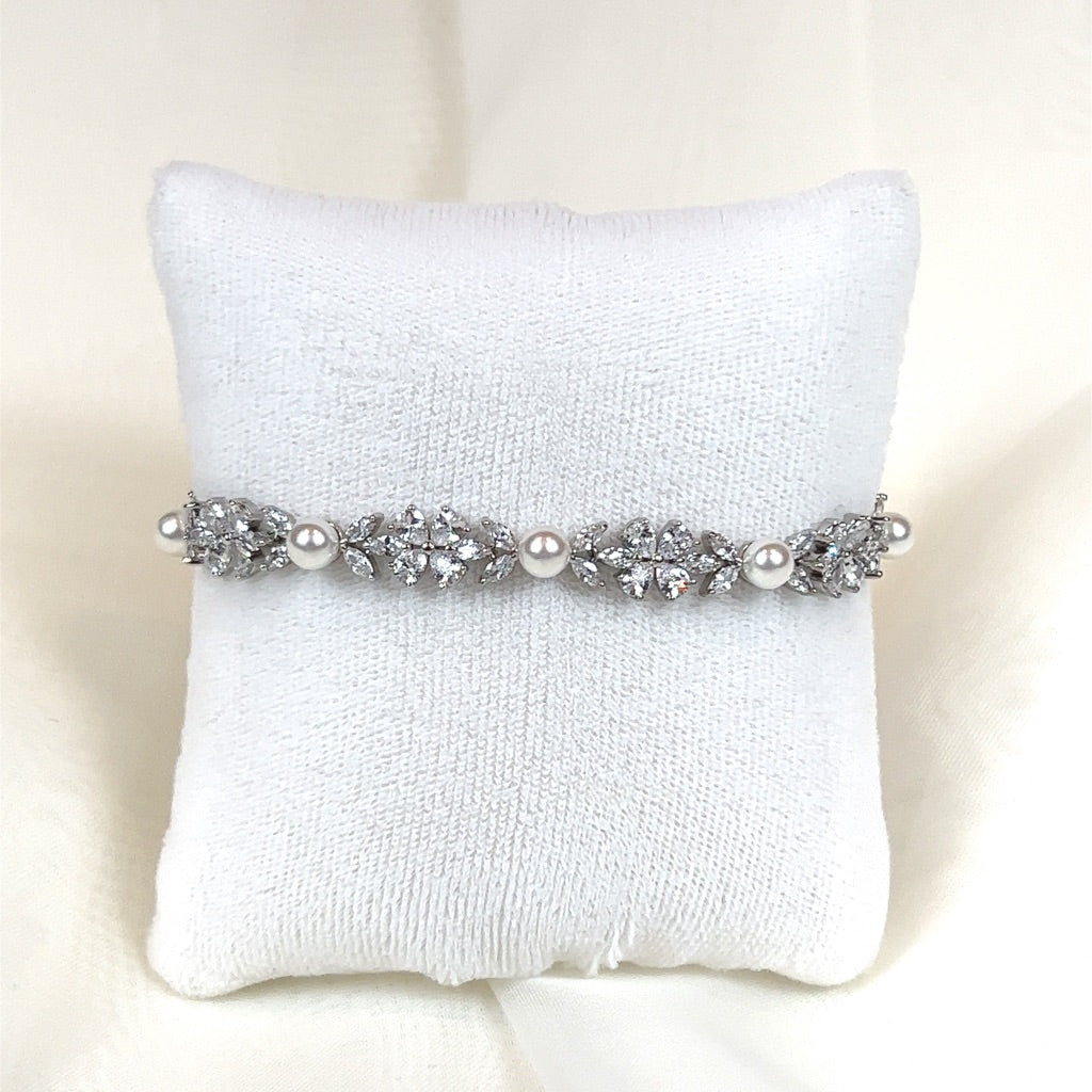 The Dainty Pearl and CZ Bracelet (style no. B147) with intricate floral designs and pearl accents is showcased on a white cushion, reminiscent of a Swarovski wedding crown, with sparkling crystal-like embellishments nestled between the pearls.
