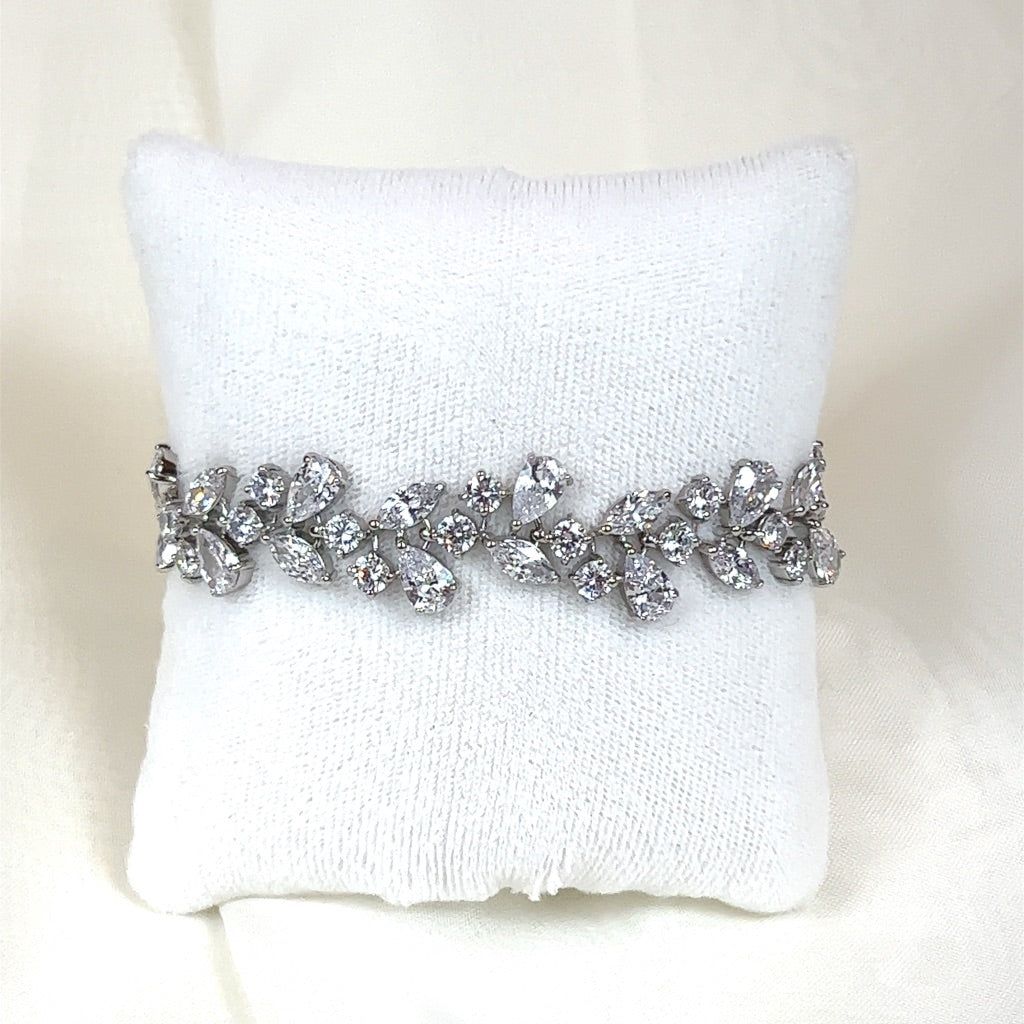 The Shimmering Cluster Bridal Bracelet (style no. B152), a silver piece adorned with CZ gemstones in a leaf pattern, rests on a white fabric cushion, radiating elegance akin to a Swarovski bridal tiara against a soft white background.