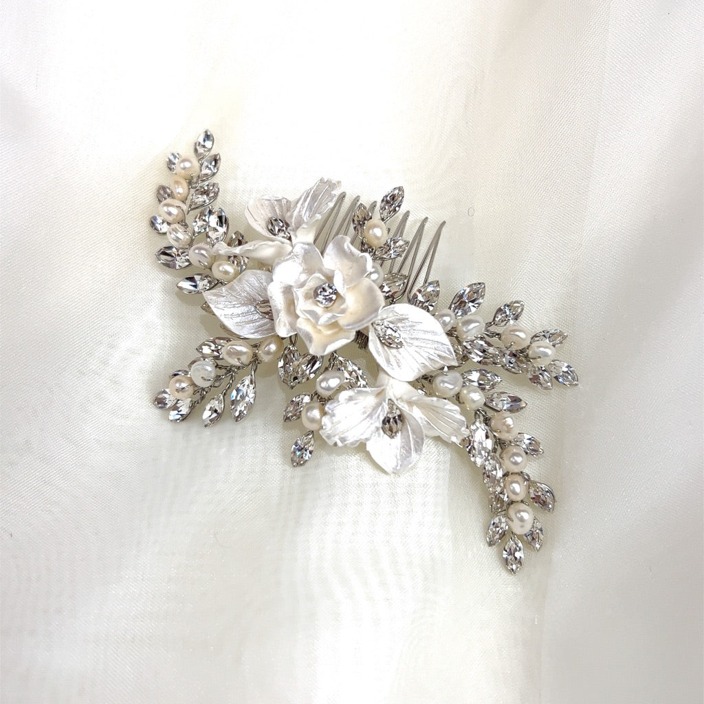The Delicate Blossoming Bridal Hair Comb (style no. 242733) showcases a decorative white flower with silver leaves, adorned with pearls and sparkling Swarovski crystals, set against a soft, light-colored fabric background.