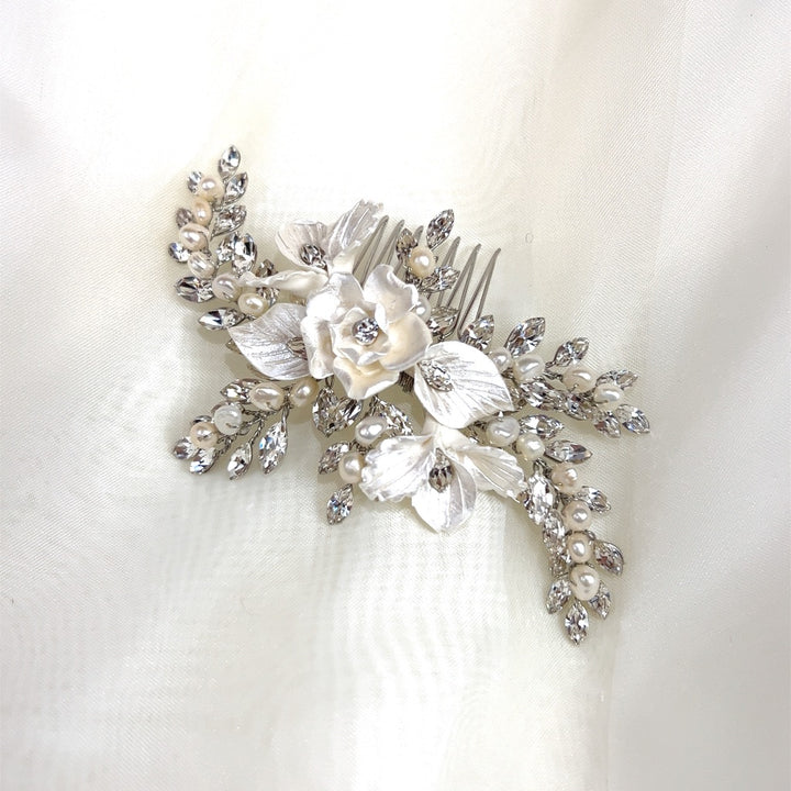The Delicate Blossoming Bridal Hair Comb (style no. 242733) showcases a decorative white flower with silver leaves, adorned with pearls and sparkling Swarovski crystals, set against a soft, light-colored fabric background.