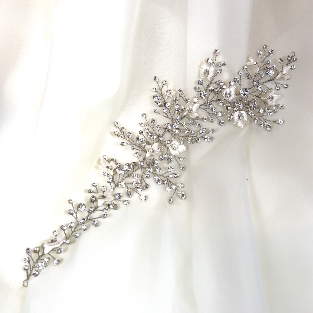The Crystal and Flower Branch Vine, style no. 242736, is a delicate Swarovski crystal bridal hair accessory featuring floral and leaf designs with sparkling crystals. It is elegantly displayed on a soft white fabric background.