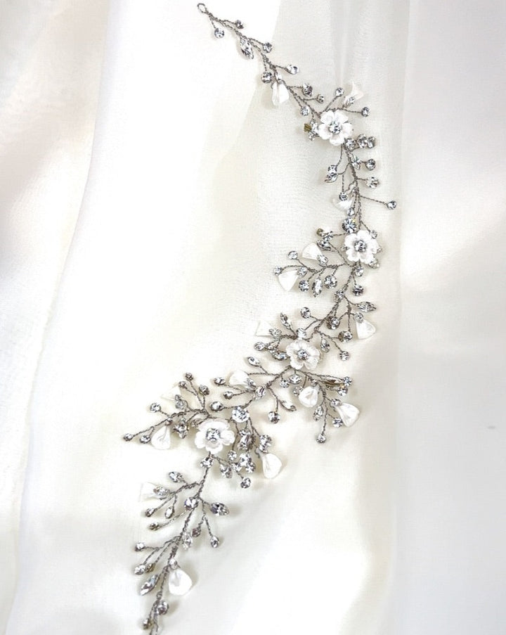 This elegant Asymmetrical Blossom Wedding Hair Vine (style no. 242735) is a delicate floral Swarovski crystal hair accessory. It features intricate details with small flowers, leaves, and sparkling crystals in a winding design, perfect for bridal elegance on white fabric.