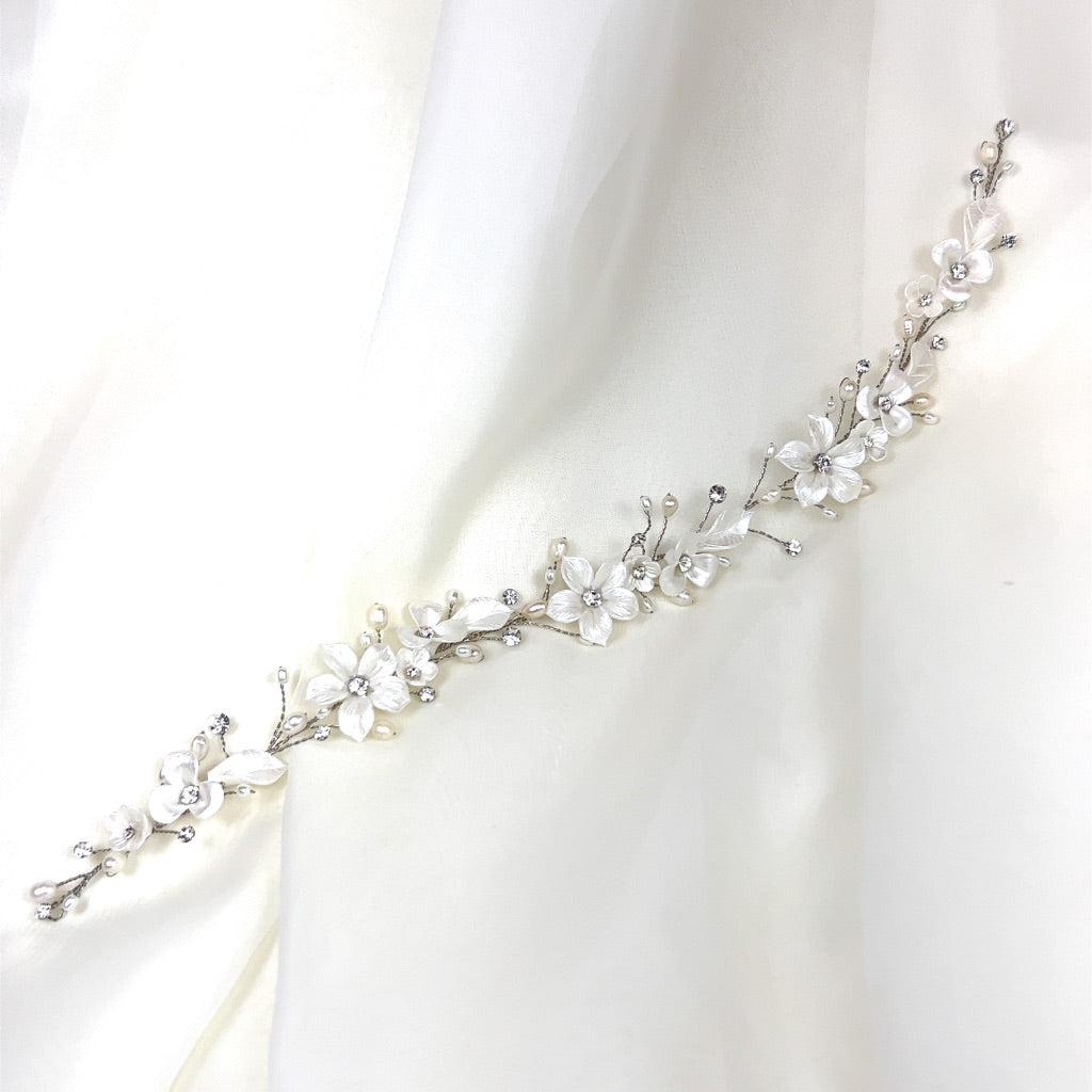 The Delicate Porcelain Flower Vine (style no. 242732) features a Swarovski headband with white flowers and sparkling gems on a silver vine, set against sheer white fabric.