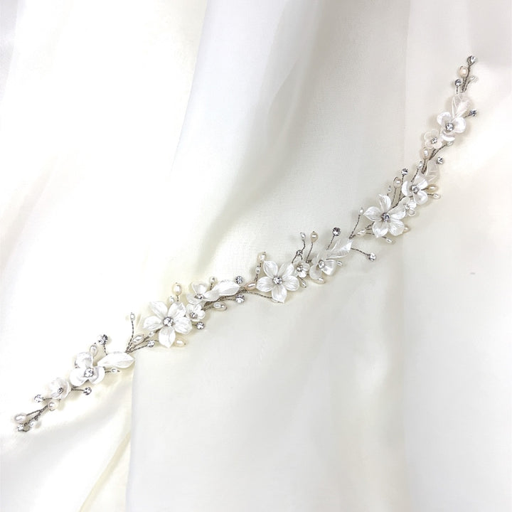 The Delicate Porcelain Flower Vine (style no. 242732) features a Swarovski headband with white flowers and sparkling gems on a silver vine, set against sheer white fabric.