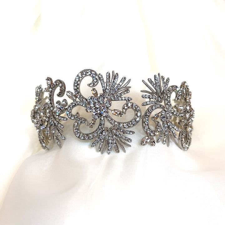 The Showstopper Crystal Headband (Style no. 3913542) features ornate silver with intricate floral and swirl designs, sparkling crystals akin to a Swarovski hair vine, displayed on a soft white fabric background.