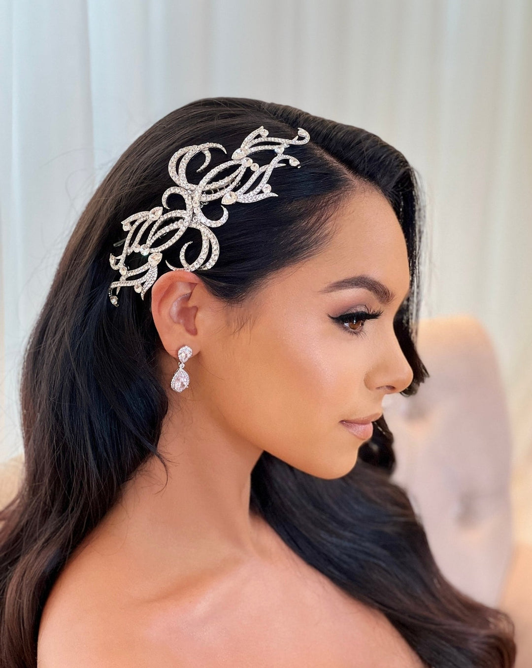 A woman with long, dark, wavy hair wears the Elegant Crystal Swirl  Bridal Wedding Comb (style no. 242634) adorned with Swarovski crystals. She has drop earrings and subtle makeup with winged eyeliner and nude lipstick, gazing sideways against a soft, light background.