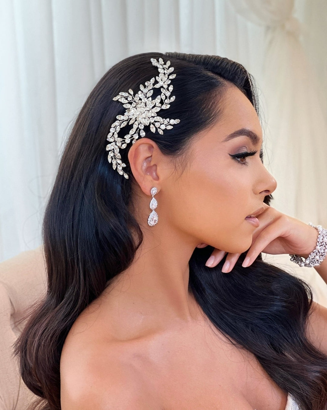 A woman with long black hair is adorned with a Delicate Flowing Swarovski Crystal Hair Comb style no. 242603, wearing dangling CZ bridal earrings and a CZ wedding bracelet. She sits in profile, resting her chin on her hand against a softly blurred background that enhances her elegance.