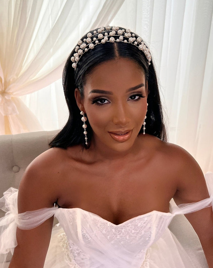 The bride, wearing an elegant white wedding dress and Swarovski bridal tiara, sits gracefully on a chair. Her hair is adorned with the Tiered Pearl Headband (Style no. 80096), complemented by pearl drop earrings. A soft curtain backdrop enhances the serene setting.