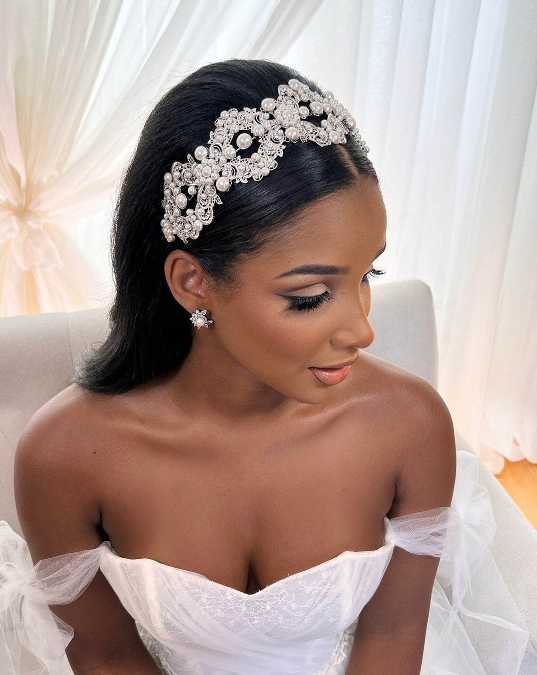 A woman in a white wedding dress sits on a light chair, her long smooth hair complemented by the Glamorous Pearl and Crystal Headband Style no. 392591. She wears matching bejeweled tiara and earrings, while soft sheer white curtains adorn the background.