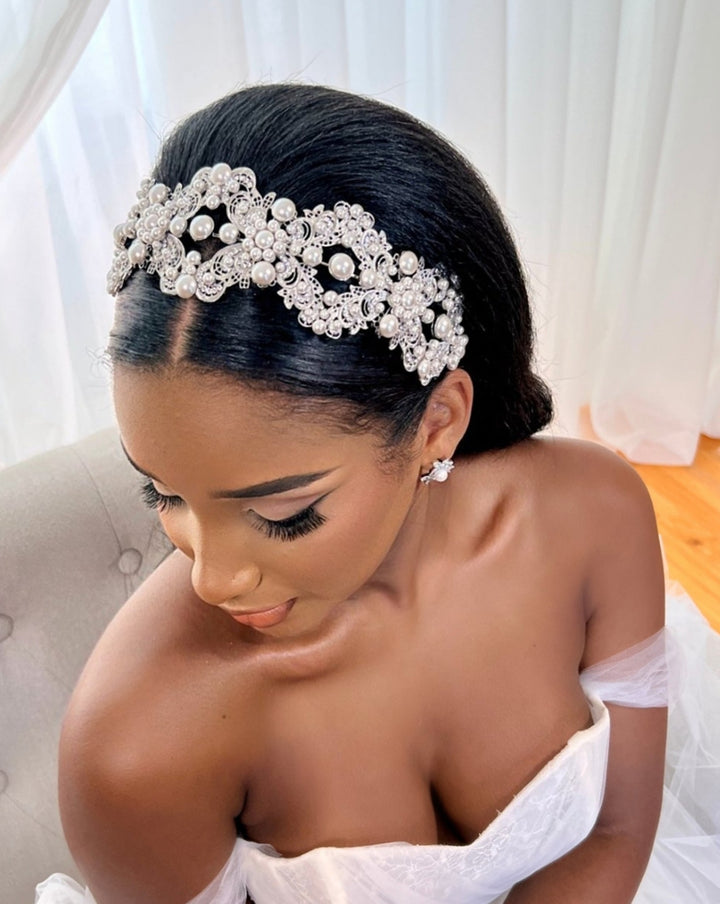 A woman sits on a chair wearing a white off-shoulder gown. Her hair is styled smooth, highlighted by the Glamorous Pearl and Crystal Wedding Headband Style no. 392591. With elegant makeup and bold eyeliner, she looks down in front of white curtains.
