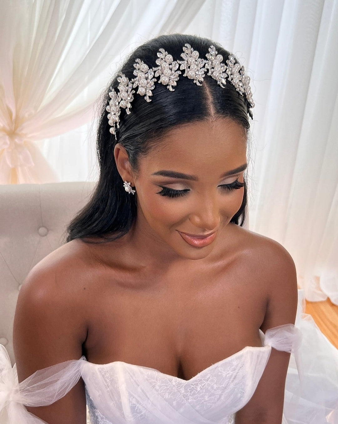 A woman in a strapless white dress sits gracefully before sheer curtains, wearing a Luxurious Pearl and Crystal Headband, Style no.80066, and matching pearl earrings. Her elegant makeup complements her cascading long hair and closed eyes, radiating contentment.
