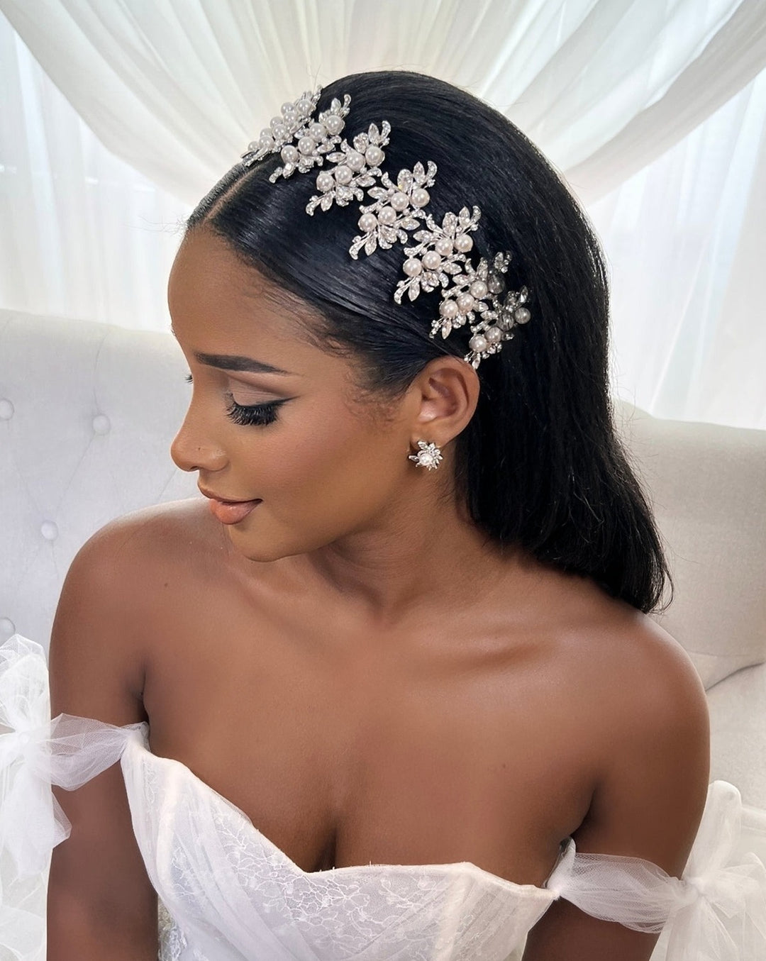 A bride in a white off-the-shoulder gown sits gracefully, adorned with a Luxurious Pearl and Swarovski Crystal Bridal Headband Style no.80066 and matching earrings, against a soft white curtain backdrop. Her long, dark hair cascades smoothly down her back.