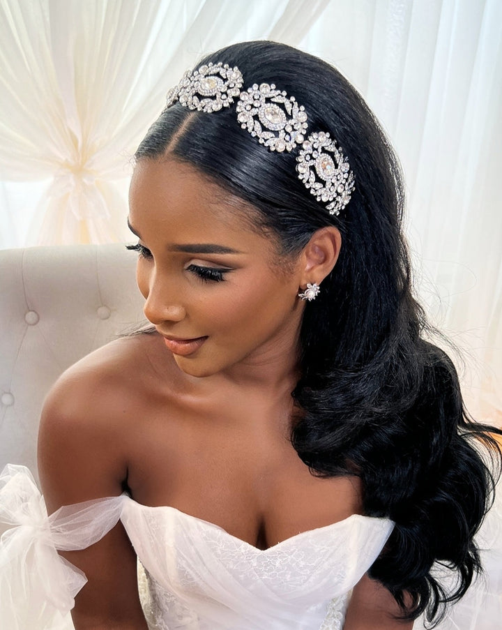 A woman in a white off-shoulder bridal gown sits on a tufted chair, her long wavy black hair adorned with the Ornate Crystal Bridal Headband Style no. 242737, matching floral earrings, soft makeup, and sheer curtains behind her.