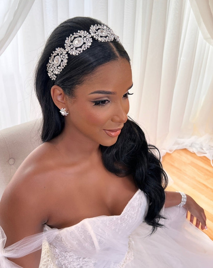A woman in a white, off-shoulder bridal gown with long, wavy black hair is seated and smiling. She wears the Ornate Crystal Bridal Headband Style no. 242737 and matching earrings against an elegant backdrop of white curtains and soft light.