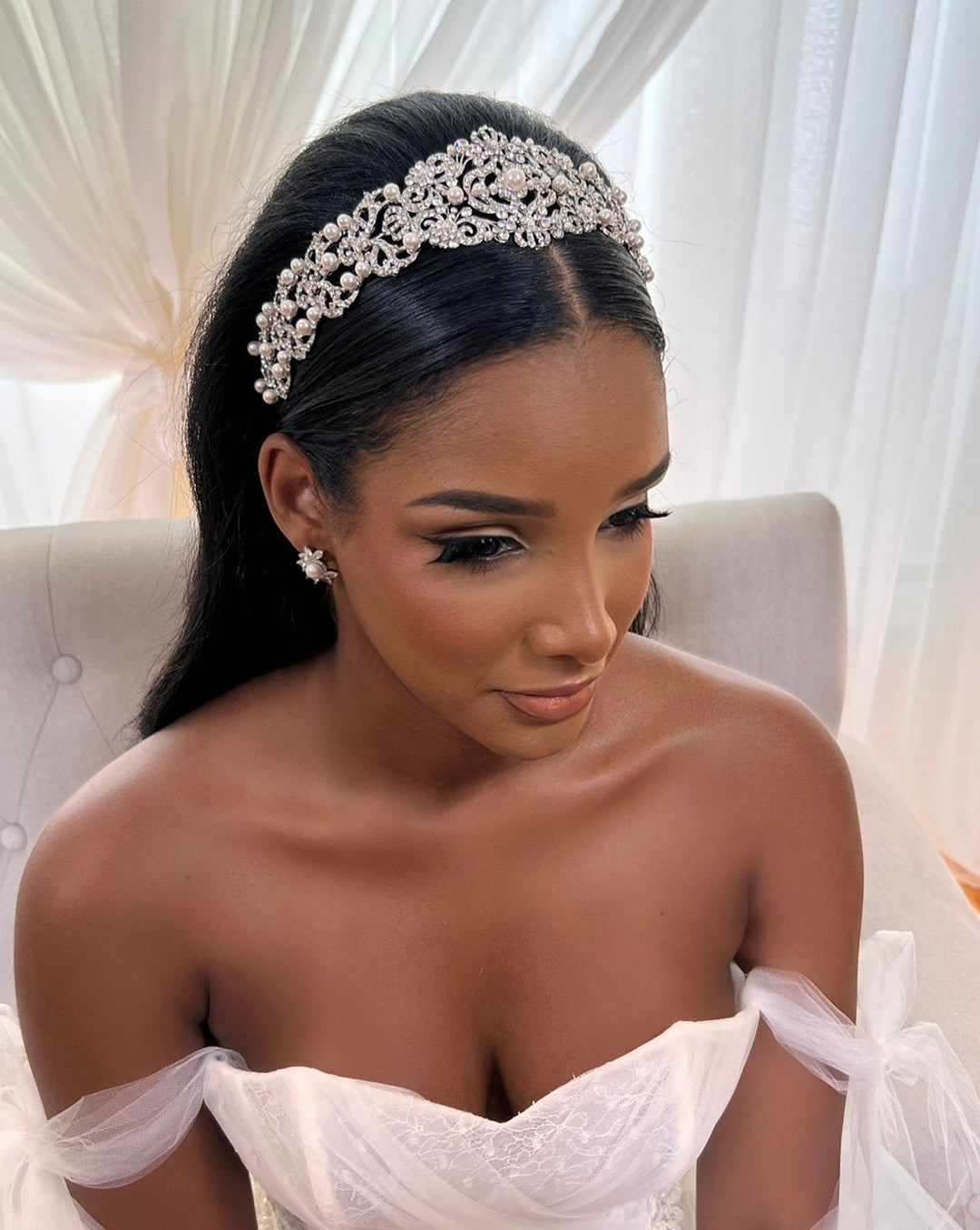 A woman sits gracefully in a white off-shoulder dress, her long dark hair cascading down. She wears a Vintage Inspired Swarovski Crystal Headband, style no. 80107, that glistens alongside her flower-shaped CZ wedding earrings earrings, while soft light through sheer curtains enhances her elegant aura.